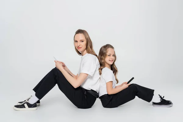 Donna e bambino con i telefoni cellulari seduti dietro e guardando la fotocamera su sfondo grigio — Foto stock