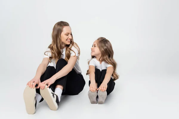 Madre e hija en ropa blanca y negra sonriéndose mientras están sentadas sobre un fondo gris - foto de stock