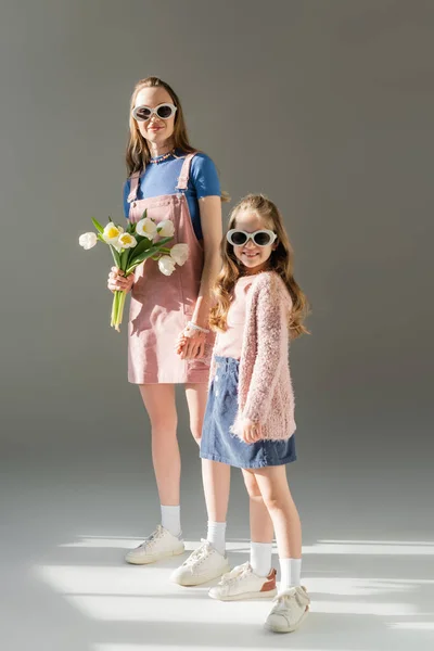 In voller Länge glückliche Mutter mit Blumen und Tochter in Sonnenbrille, Händchen haltend auf grau — Stockfoto