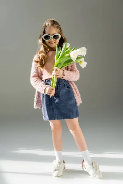 Pleine longueur de gosse heureux dans des lunettes de soleil tenant des fleurs sur le gris — Photo de stock