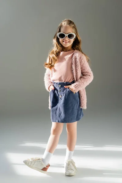 Full length of happy kid in sunglasses standing with hands in pockets on grey — Stock Photo