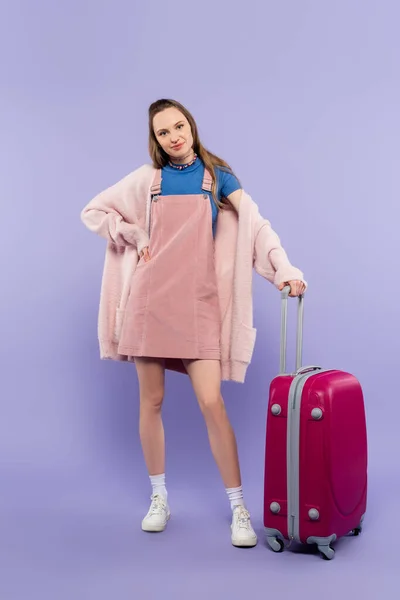 Full length of woman in pink overall dress standing with hand on hip near luggage on purple — Stock Photo