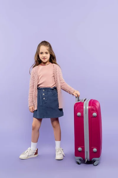 Повна довжина веселої дівчини, що стоїть з рожевим багажем на фіолетовому — стокове фото