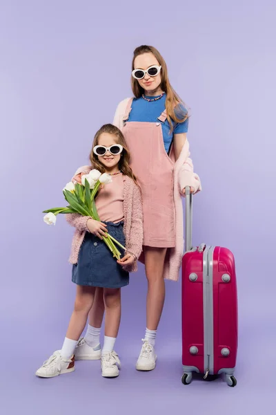 Повна довжина матері і дитини в сонцезахисних окулярах, що тримають квіти і стоять біля багажу на фіолетовому — стокове фото