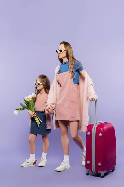 Mutter und Kind in Sonnenbrille halten sich an Händen und gehen mit Gepäck auf lila — Stockfoto