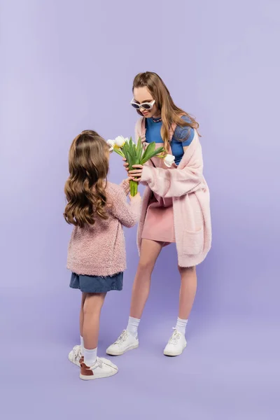 Kind in voller Länge mit Sonnenbrille schenkt Mutter Blumen im Ganzkörperkleid auf lila — Stockfoto