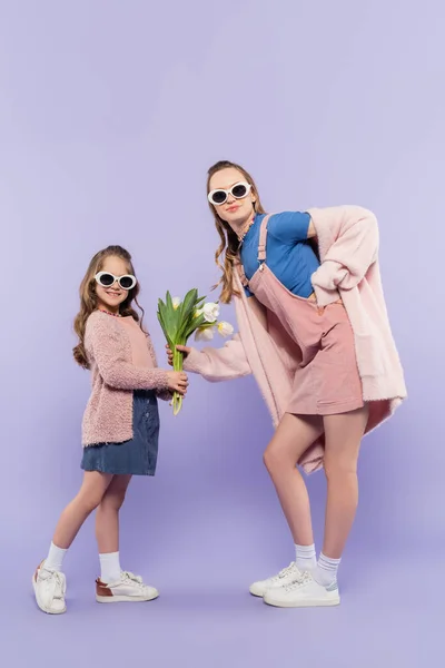 Счастливая девочка в солнцезащитных очках в полный рост дарит цветы маме в платье фиолетового цвета — стоковое фото