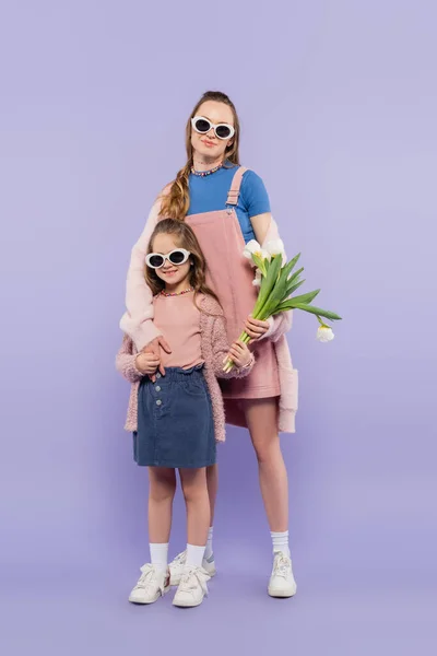 Kind in voller Länge mit Sonnenbrille und Blumen in der Nähe der Mutter im Kleid auf lila — Stockfoto