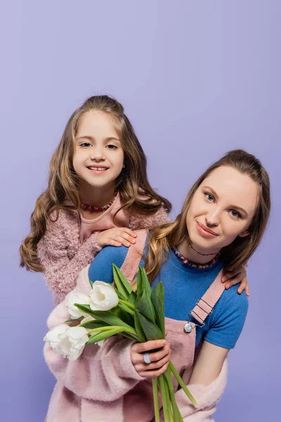 Femme souriante tenant des tulipes près de fille heureuse isolée sur violet — Photo de stock