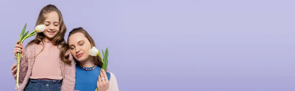Довольные мать и дочь с закрытыми глазами держа тюльпаны изолированы на фиолетовый, баннер — стоковое фото