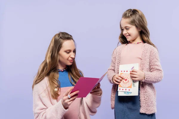 Щаслива мати читає вітальні листівки біля веселої дочки ізольовані на фіолетовому, концепція Дня матері — стокове фото