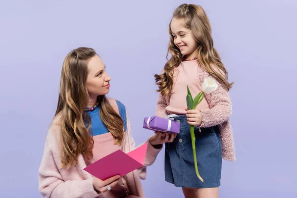 Menina feliz segurando caixa de presente e tulipa perto da mãe com cartão de saudação isolado em roxo — Fotografia de Stock