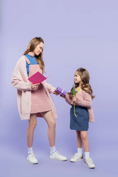 Comprimento total da menina feliz segurando caixa de presente e tulipa perto da mãe com cartão de saudação no roxo — Fotografia de Stock