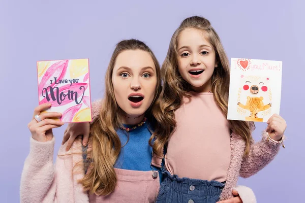 Madre e hija asombradas sosteniendo tarjetas de felicitación aisladas en púrpura, concepto del día de las madres - foto de stock