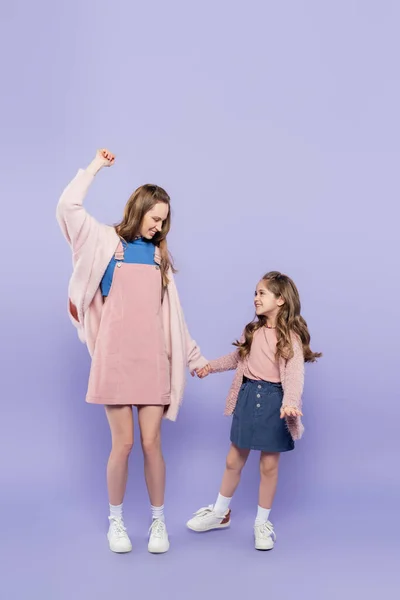 Full length of happy mother in pink overall dress holding hands with joyful girl on purple — Stock Photo