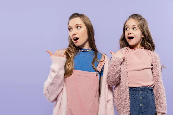 Mãe espantada e filha apontando com os polegares e olhando para longe isolado em roxo — Fotografia de Stock