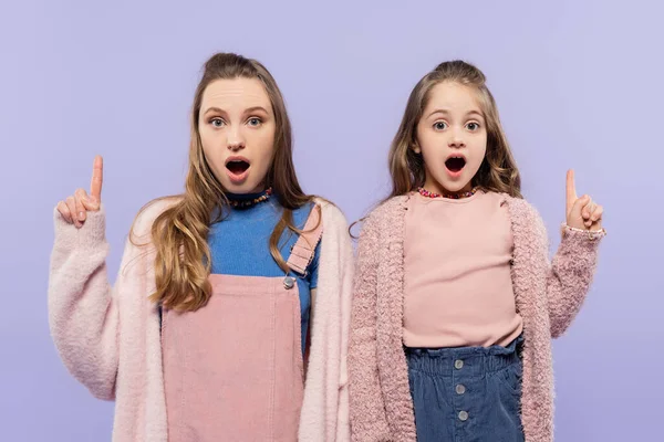 Chocado mãe e filha apontando com os dedos para cima e olhando para a câmera isolada em roxo — Fotografia de Stock