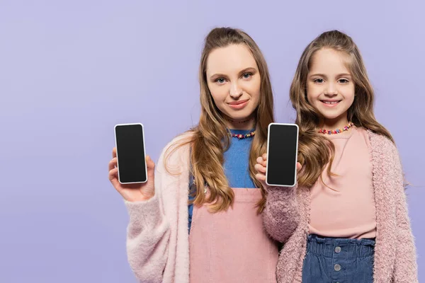 Счастливая мать и дочь показывая смартфоны с чистым экраном изолированы на фиолетовый — стоковое фото