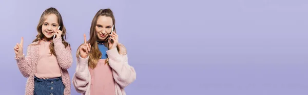 Feliz mãe e filha falando em telefones celulares e apontando para cima isolado em roxo, banner — Fotografia de Stock