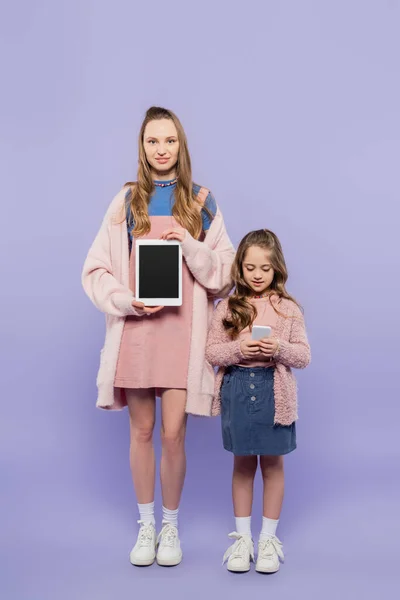 Longitud completa de la mujer que muestra la tableta digital con la pantalla en blanco cerca del niño usando el teléfono inteligente en púrpura - foto de stock