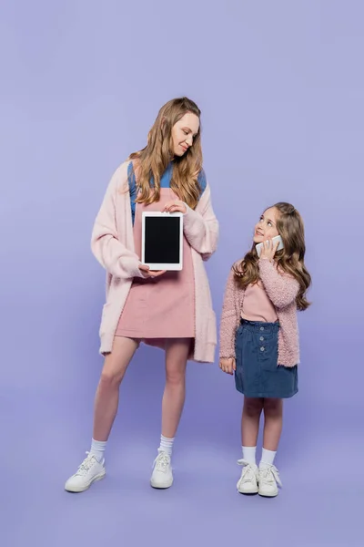 Volle Länge der Frau zeigt digitale Tablette mit leerem Bildschirm in der Nähe von Kind spricht auf Smartphone auf lila — Stockfoto