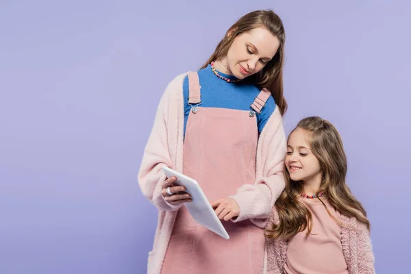 Donna sorridente mostrando tablet digitale a figlia felice isolato su viola — Foto stock