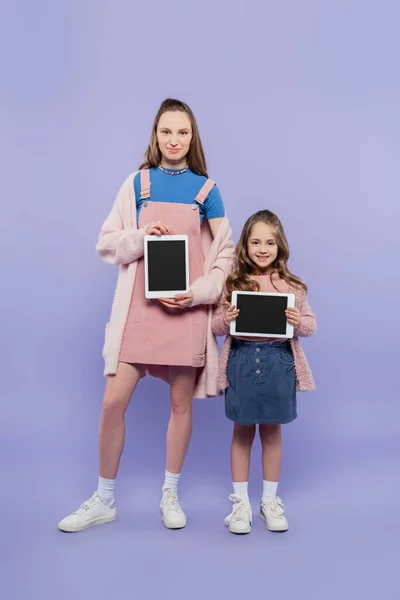 Longitud completa de madre e hija sosteniendo tabletas digitales con pantalla en blanco en púrpura - foto de stock