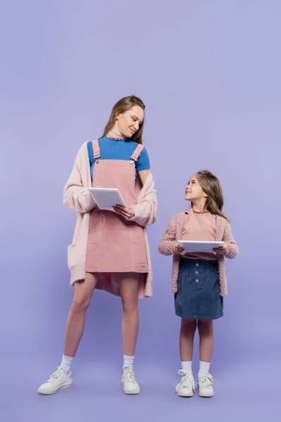 Piena lunghezza di madre e figlia che tengono in mano compresse digitali mentre si guardano l'un l'altro su viola — Foto stock