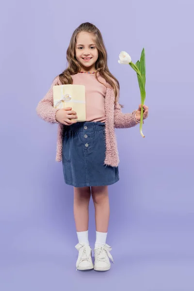 Pleine longueur de fille heureuse tenant boîte cadeau et tulipe sur violet — Photo de stock