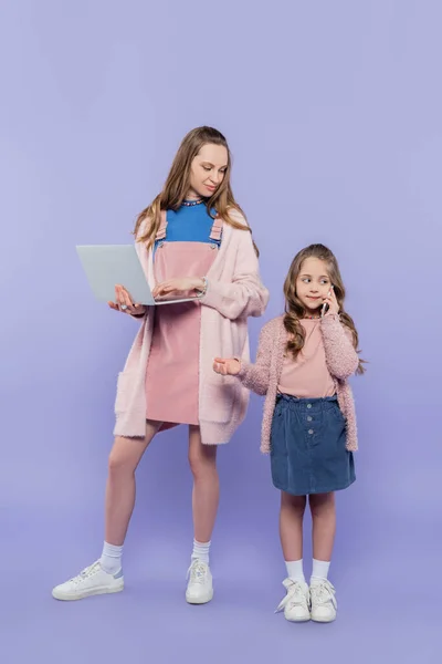 Full length of mother with laptop looking at child talking on smartphone on purple — Stock Photo