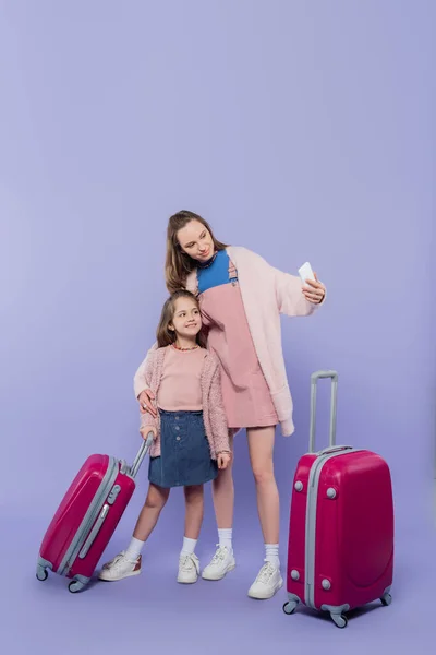 Longitud completa de la madre feliz y el niño tomando selfie cerca de equipaje en púrpura - foto de stock