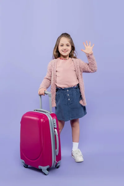 Comprimento total da menina feliz acenando mão perto de bagagem rosa em roxo — Fotografia de Stock