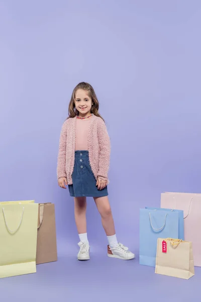 Longitud completa de niño feliz de pie cerca de bolsas de compras en púrpura - foto de stock