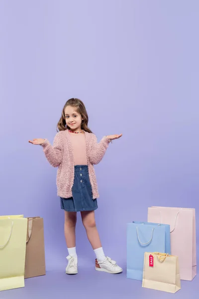 Comprimento total da criança feliz apontando com as mãos perto de sacos de compras em roxo — Fotografia de Stock