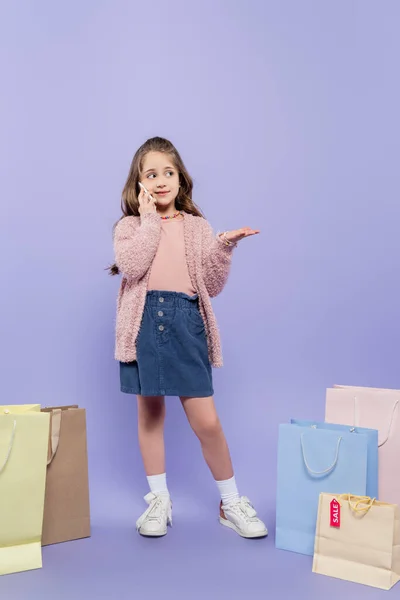 Full length of happy kid talking on smartphone and standing near shopping bags on purple — Stock Photo