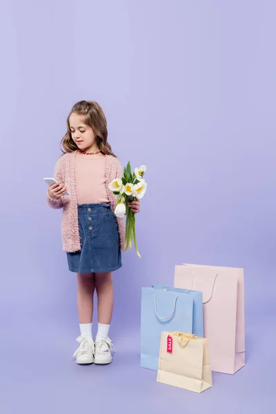 Повна довжина дитини, що тримає квіти і використовує смартфон, стоячи біля сумки для покупок на фіолетовому — стокове фото