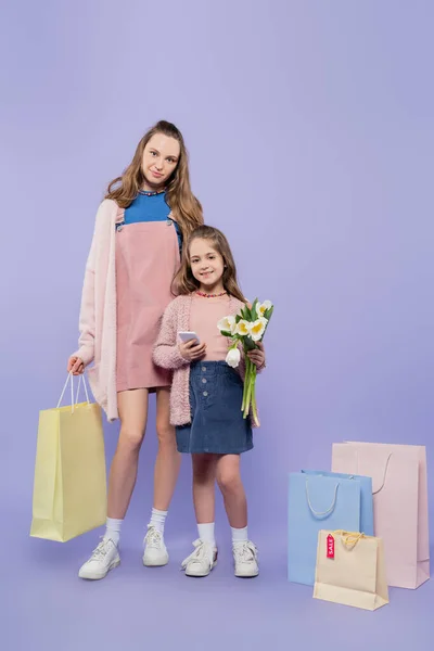 Full length of kid holding flowers and using smartphone while standing with mother near shopping bags on purple — Stock Photo