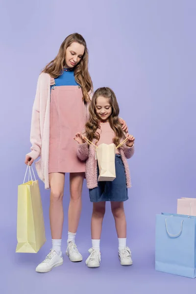 Full length of kid looking inside of shopping bag near mother on purple — Stock Photo