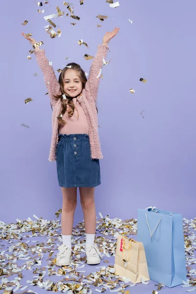 Longitud completa de la chica feliz de pie cerca de la caída de confeti y bolsas de compras en púrpura - foto de stock