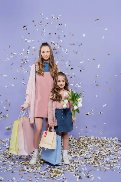 Pleine longueur de mère heureuse et fille tenant des sacs à provisions et des fleurs près de confettis tombant sur violet — Photo de stock