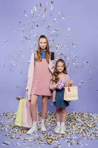 Comprimento total da mãe feliz segurando smartphone e sacos de compras perto da filha com presente quase caindo confete no roxo — Fotografia de Stock