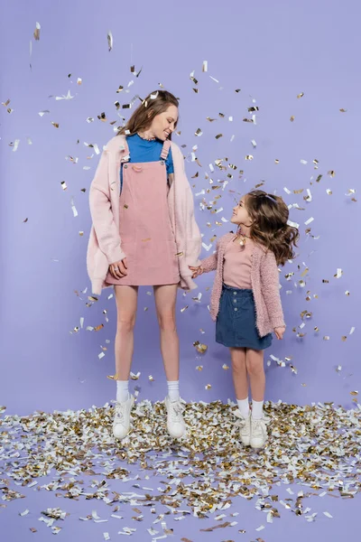 Longitud completa de feliz madre e hija tomados de la mano y saltando cerca de caer confeti en púrpura - foto de stock