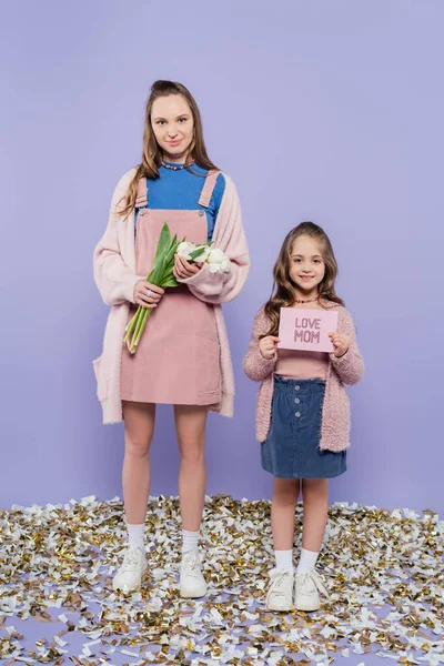 Pleine longueur de mère heureuse tenant des fleurs près de l'enfant avec carte de vœux sur la fête des mères sur violet — Photo de stock