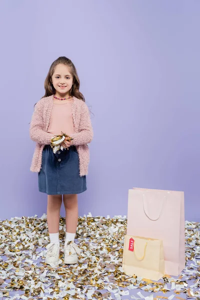 Pleine longueur de fille heureuse tenant confettis près de sacs à provisions sur violet — Photo de stock