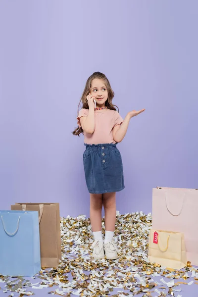 Comprimento total da menina falando no telefone celular perto de sacos de compras e gestos em roxo — Fotografia de Stock