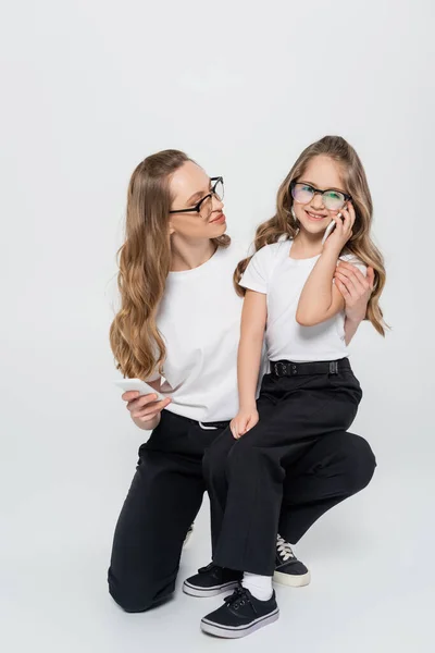 Mädchen mit Brille spricht auf Smartphone und sitzt auf dem Knie der Mutter auf grau — Stockfoto