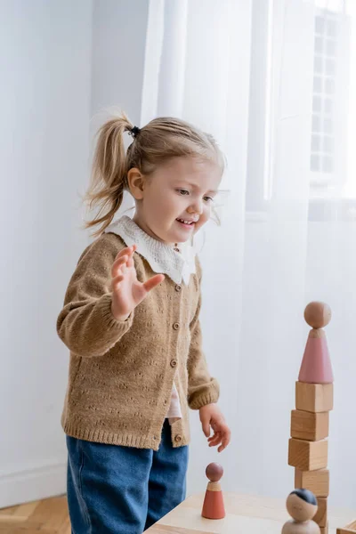 Menina feliz olhando para a torre feita de cubos de madeira e estatueta enquanto joga em casa — Fotografia de Stock