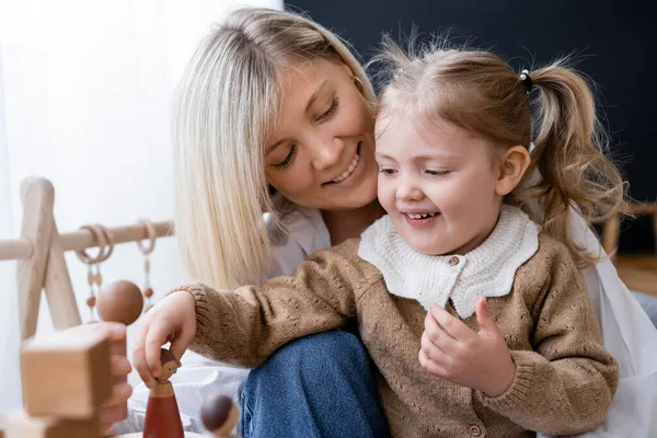 Menina feliz brincando com estatuetas de madeira perto de mãe sorridente em casa — Fotografia de Stock