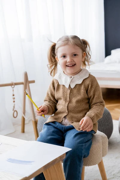 Criança alegre olhando para a câmera enquanto segurando lápis amarelo perto de papel com traços azuis — Fotografia de Stock