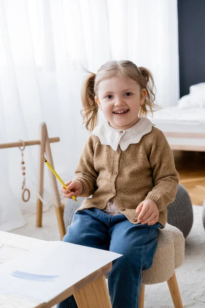 Cheerful child looking at camera while holding yellow pencil near paper with blue strokes — Stock Photo